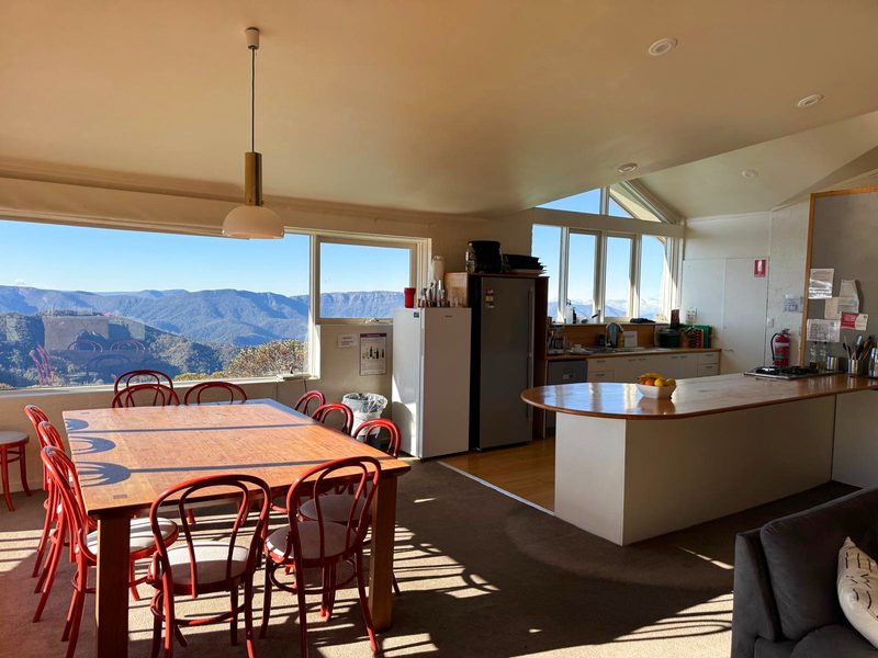 The top-level kitchen and dining: a long timber table with red bentwood chairs in the sun, the kitchen bench beyond, and windows filled with the view out across the ranges.