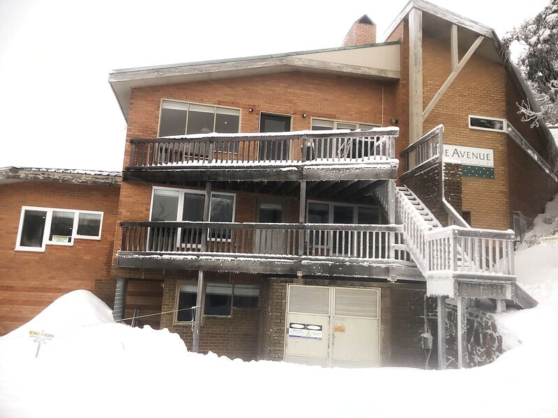 The full front of the lodge in mid-winter, balconies and external stair under snow, deep snowbanks below.